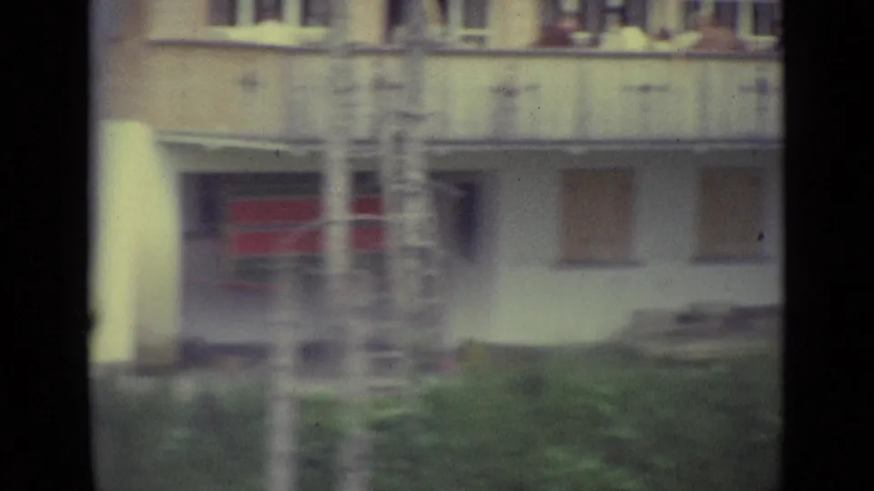 Soft 1962 Lucerne home-movie frame with the building balcony, ladder, lower red window area, and green lawn in view.