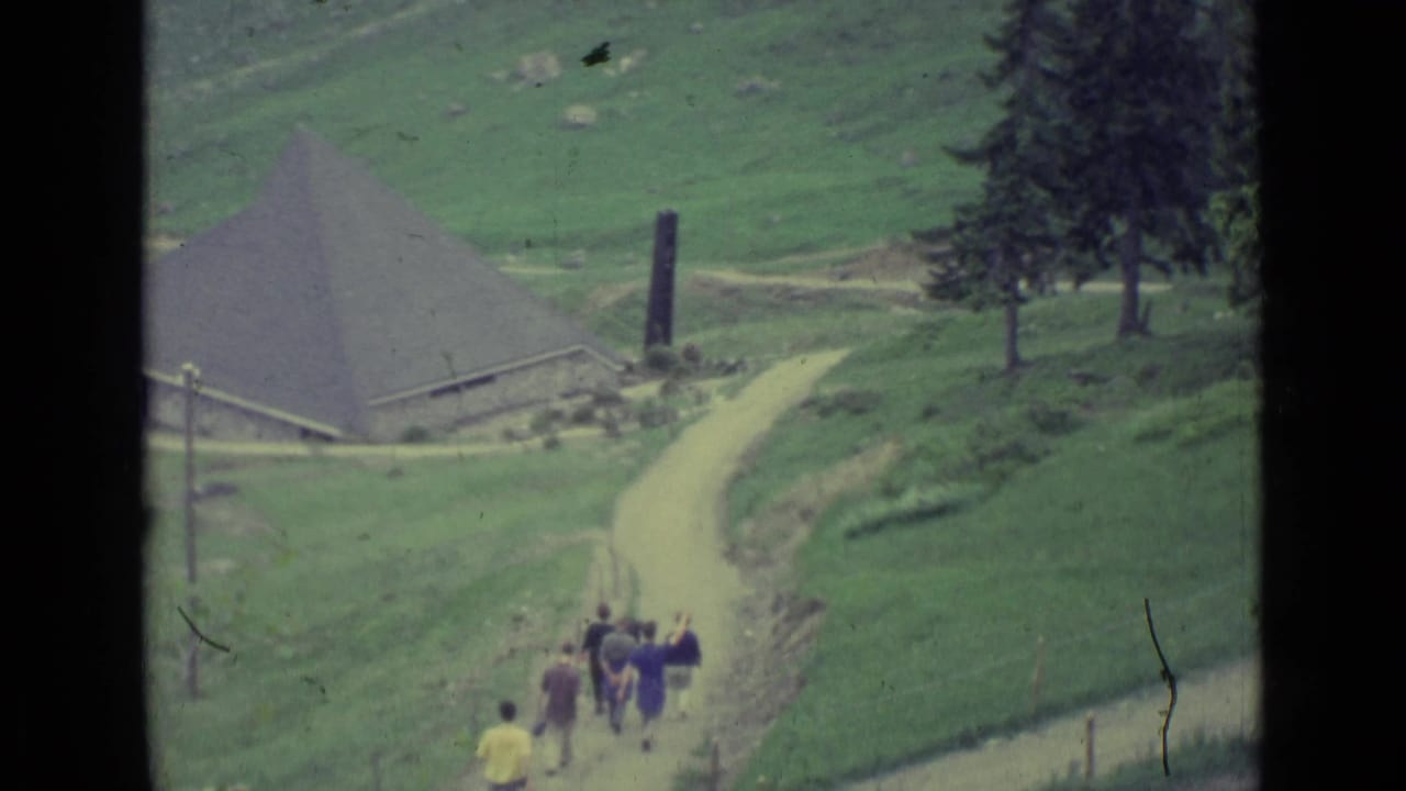 1962 Lucerne-region frame showing people walking along a winding dirt path toward a conical-roofed rural building.
