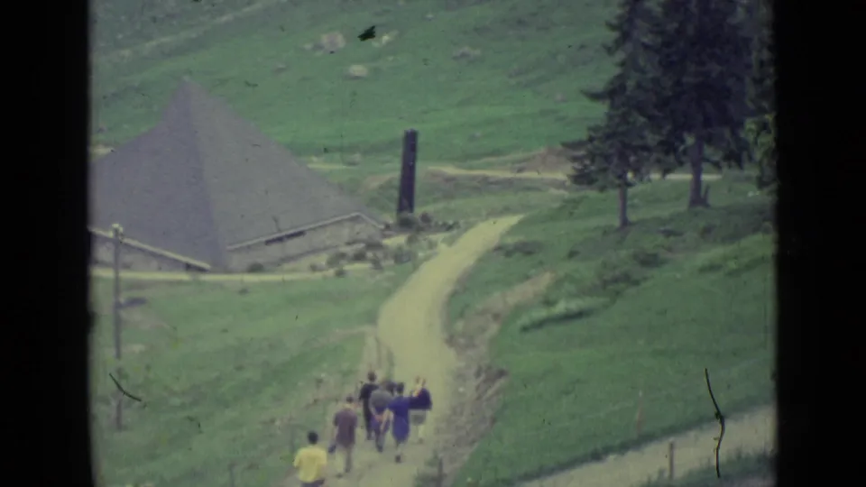1962 Lucerne-region frame showing people walking along a winding dirt path toward a conical-roofed rural building.
