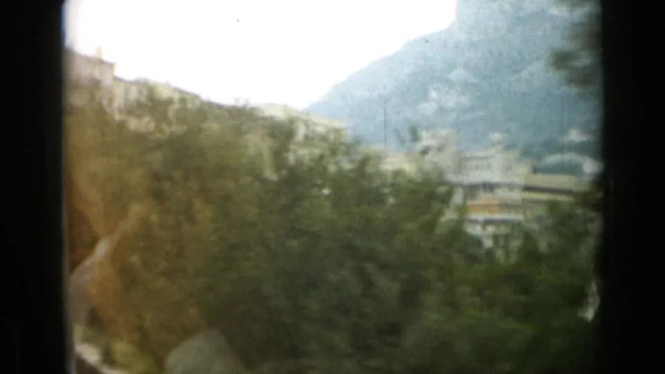 Soft 1962 Monaco view with trees in the foreground, hillside buildings in the distance, and dark frame bars from the 8mm scan.