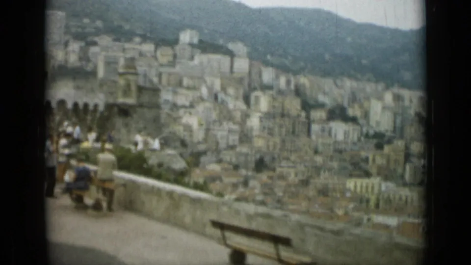 1962 Monaco overlook with visitors near a parapet, benches, crowded hillside buildings, and a mountain rising behind the city.