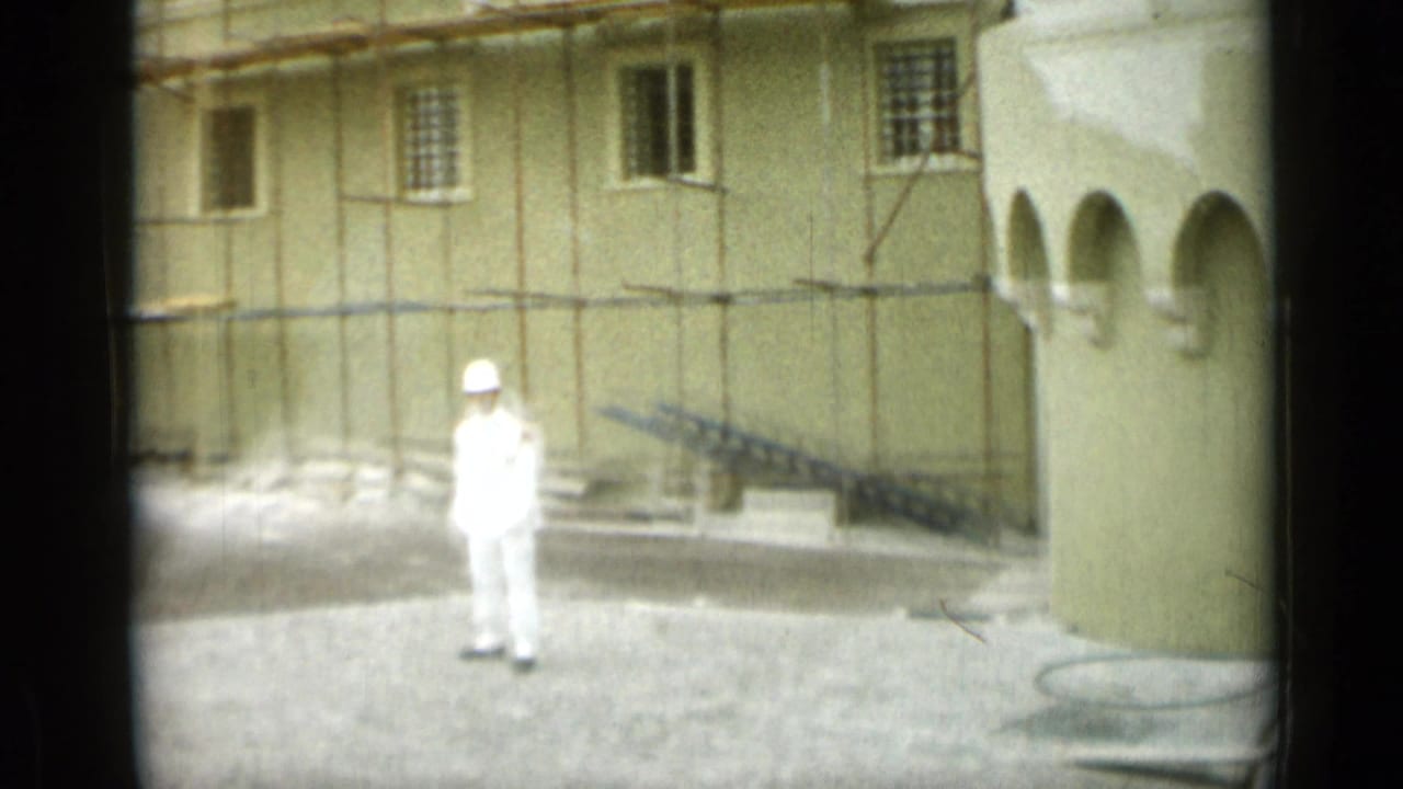 White-uniformed guard standing near a pale building with scaffolding and an arched corner.