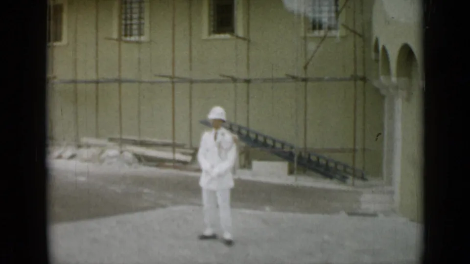 1962 Monaco 8mm frame with a guard standing still in front of scaffolding, a pale facade, barred windows, and a dark sloped railing.