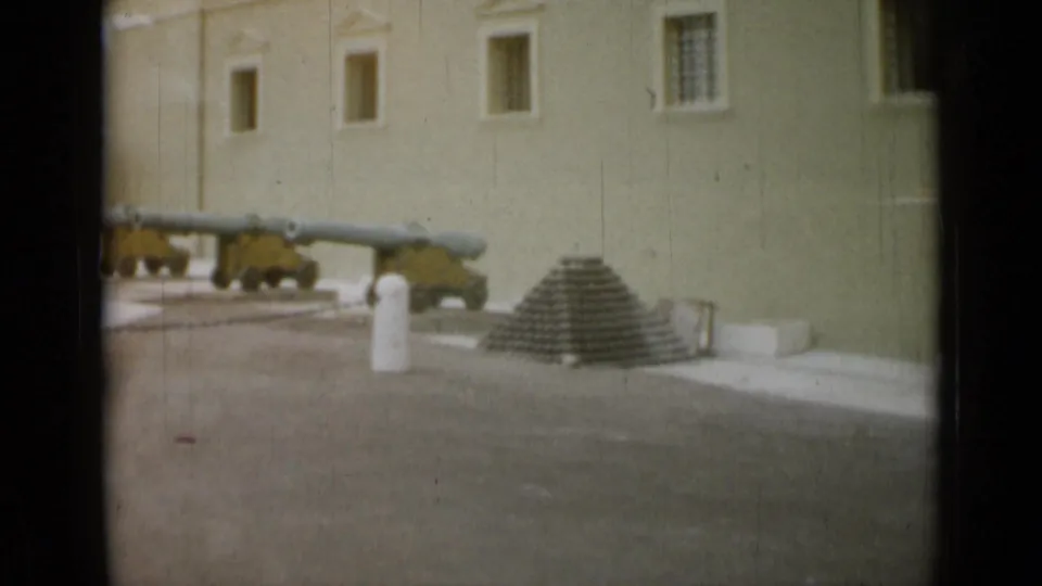 1962 Monaco 8mm frame showing three green cannons on yellow carriages beside stacked cannonballs and a pale historic building wall.