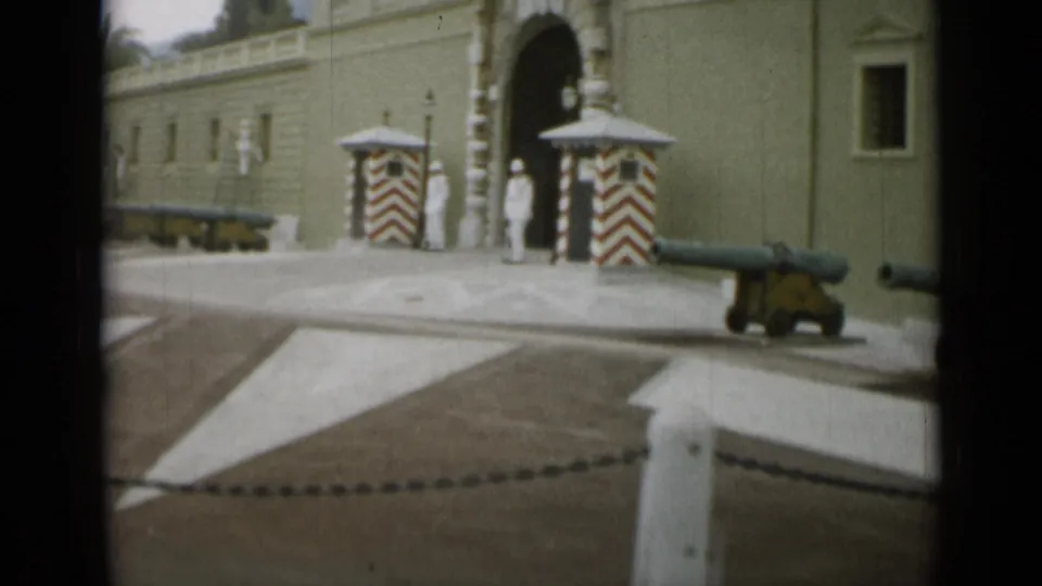 1962 Monaco frame with palace cannons in the foreground and white-uniformed guards beside red-and-white sentry boxes near the arch.