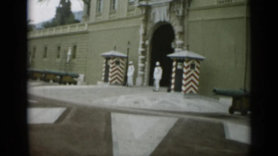 1962 Monaco home-movie view of a palace arch, guards in white, striped sentry boxes, and a row of cannons along the facade.