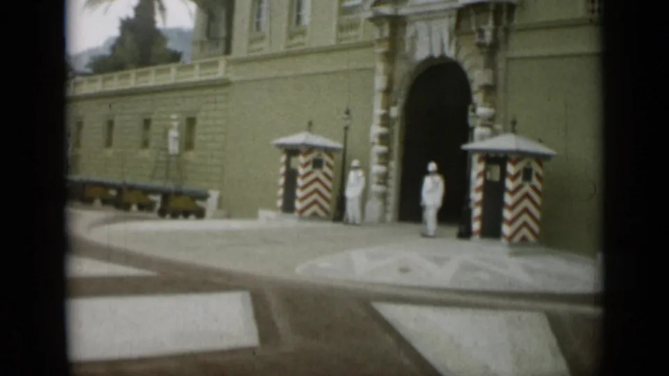 1962 Monaco frame showing a guarded palace entrance with red-and-white sentry boxes, white uniforms, cannon display, and stone paving.