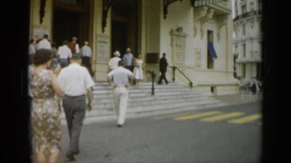 People in summer clothing walk toward the steps of a formal building in Monaco, with a uniformed guard nearby.