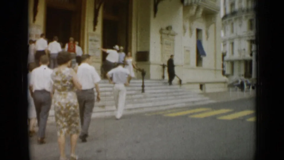 Visitors in 1960s clothing walk up the steps of a formal Monaco building as others gather near the doorway.