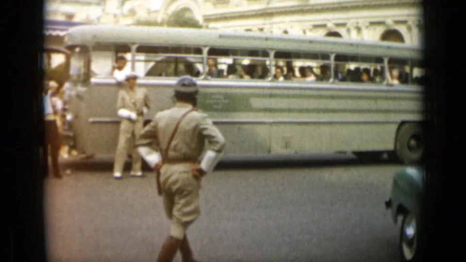 In Monaco, 1962, a helmeted uniformed man walks away as a large light-colored bus fills the background with passengers inside.