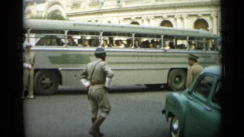 1962 Monaco street view with a uniformed officer walking near a light-colored passenger bus and a parked green car.