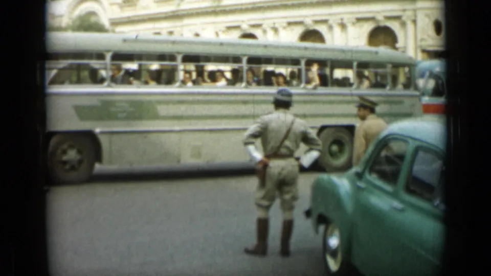 1962 Monaco street frame with a uniformed officer viewed from behind, a passenger bus, and a parked green car.
