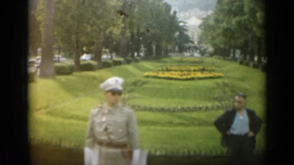 A uniformed guard in a white hat passes through a Monaco park view with lawns, flowerbeds, trees, and a man standing to the side.