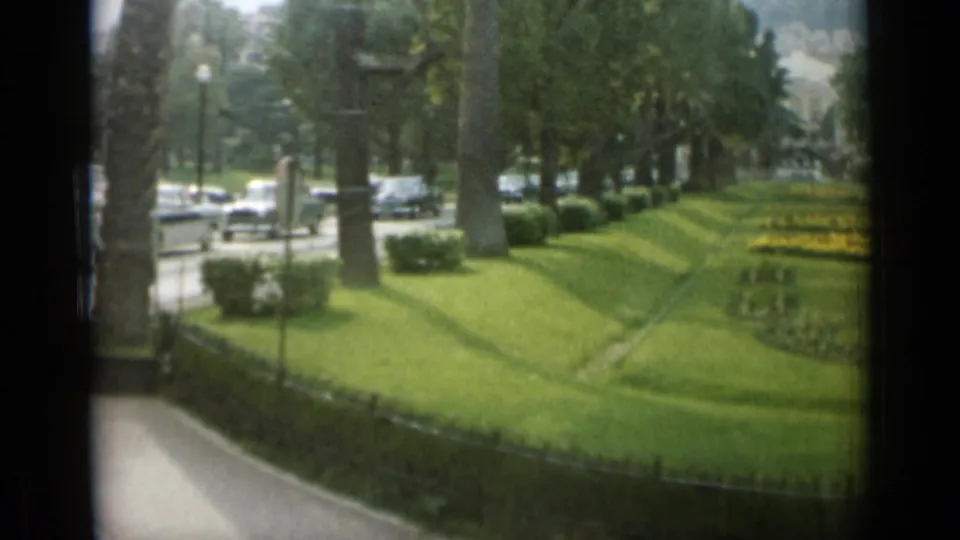 A Monaco park cutaway shows patterned grass, yellow flowers, trees, a low hedge, and period cars moving or parked along the street.