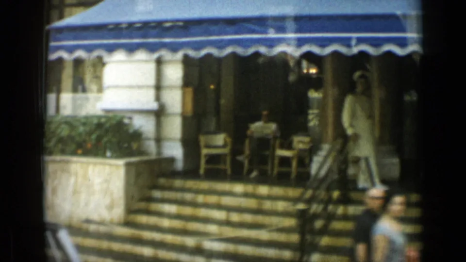 Monaco storefront with a striped blue awning, stone steps, outdoor chairs, and a uniformed person standing near the entrance.