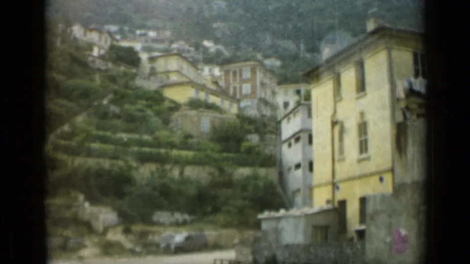 1962 Monaco home-movie frame showing a prominent yellow building, hillside houses, and green terraces beyond a roadside area.