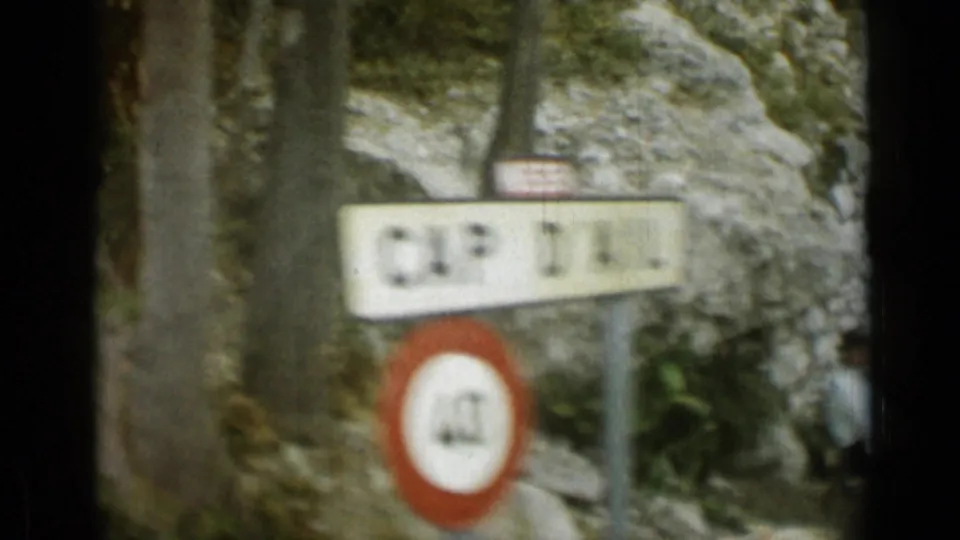 1962 Monaco frame shifting to a close roadside view with a blurred place sign, circular speed sign, rock face, and partial figure at the edge.