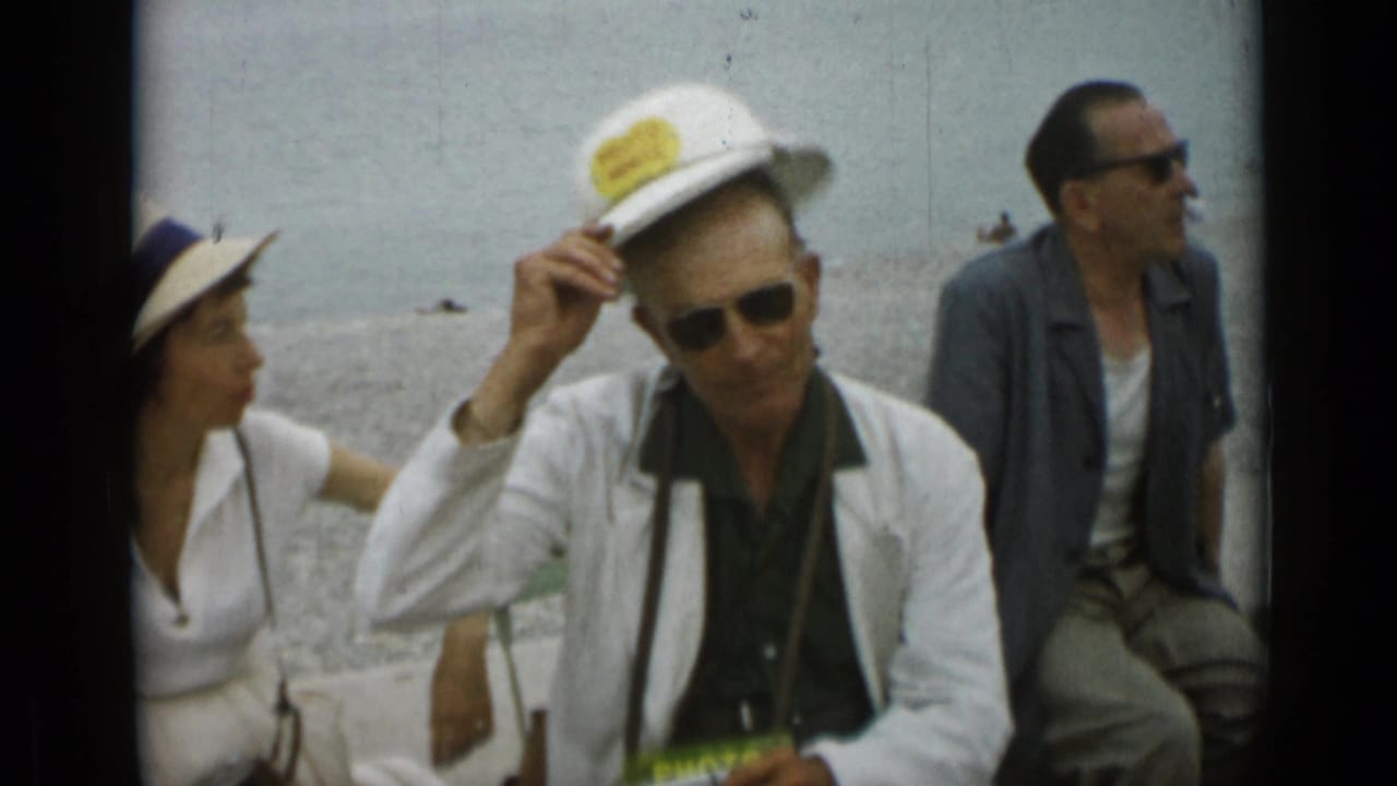 Adults seated near water, including a man adjusting a white hat and wearing sunglasses.