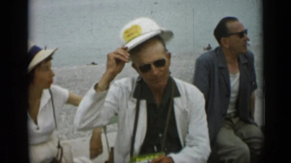 1962 Monaco home-movie frame of seated adults by the waterfront, with a man in sunglasses adjusting a white hat.