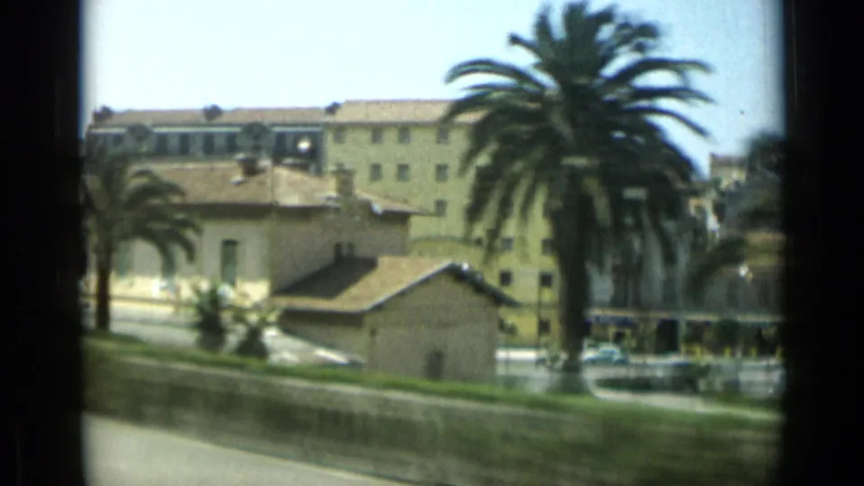 Palm-lined Monaco street view from 1962, with low buildings, a larger yellow block, cars, and greenery seen from a moving vehicle.