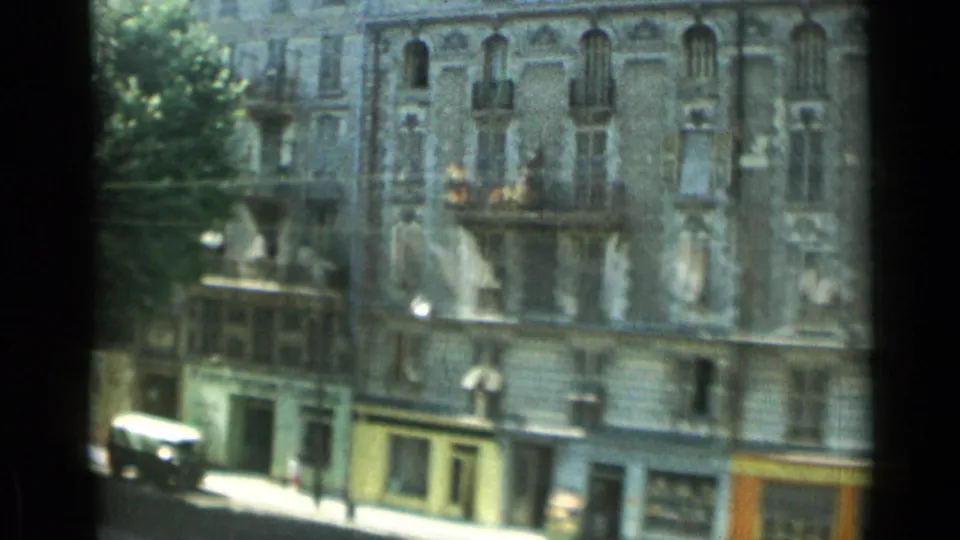 1962 Monaco street scene with an ornate stone facade, balconies, street-level shops, awnings, and a vehicle near the curb.