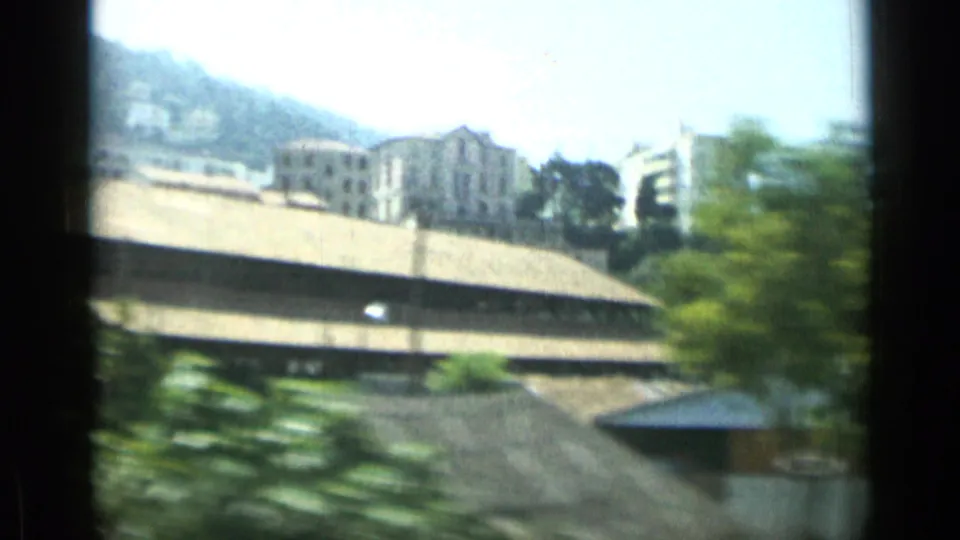 Blurred 1962 Monaco vehicle view toward hillside buildings, pale rooftops, trees, and distant apartment blocks.