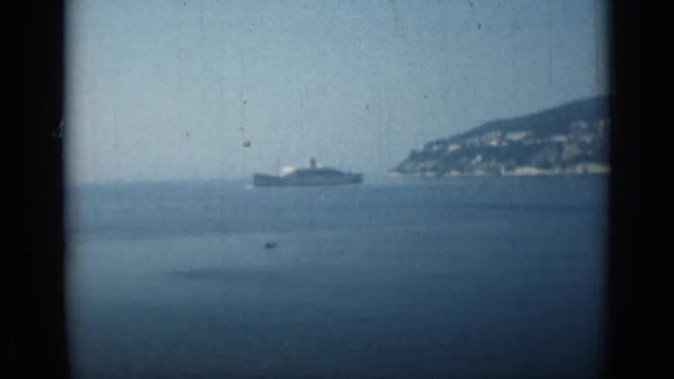 Monaco coastal home-movie frame with a distant vessel, open water, and a rocky hillside shoreline at frame right.