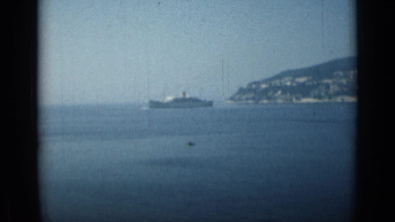 Distant passenger ship on blue water with a hilly Monaco shoreline behind it.