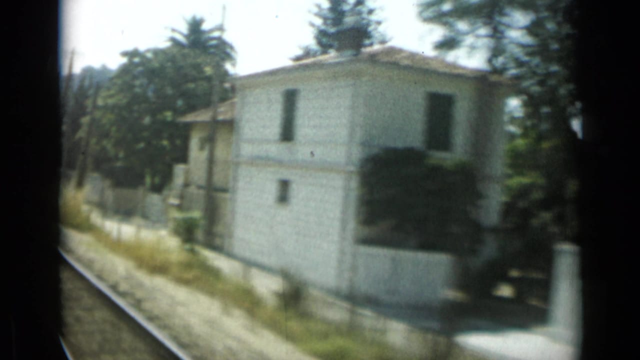 White roadside villa with shuttered windows, low wall, trees, and rail or road edge passing by.