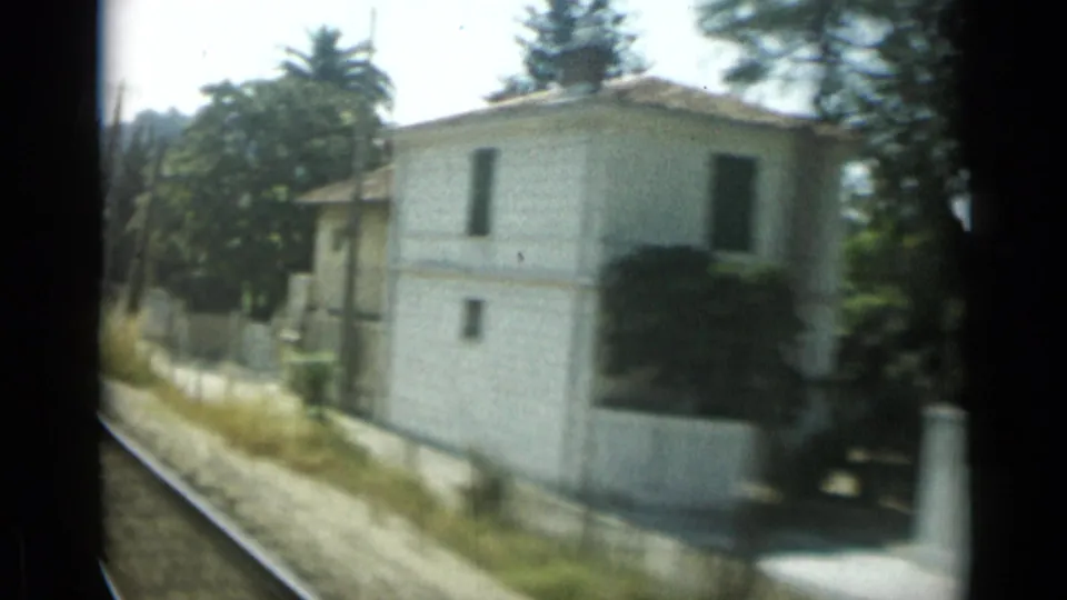 1962 Monaco home-movie view from a moving vehicle past a white villa, garden wall, roadside grass, and tall trees.