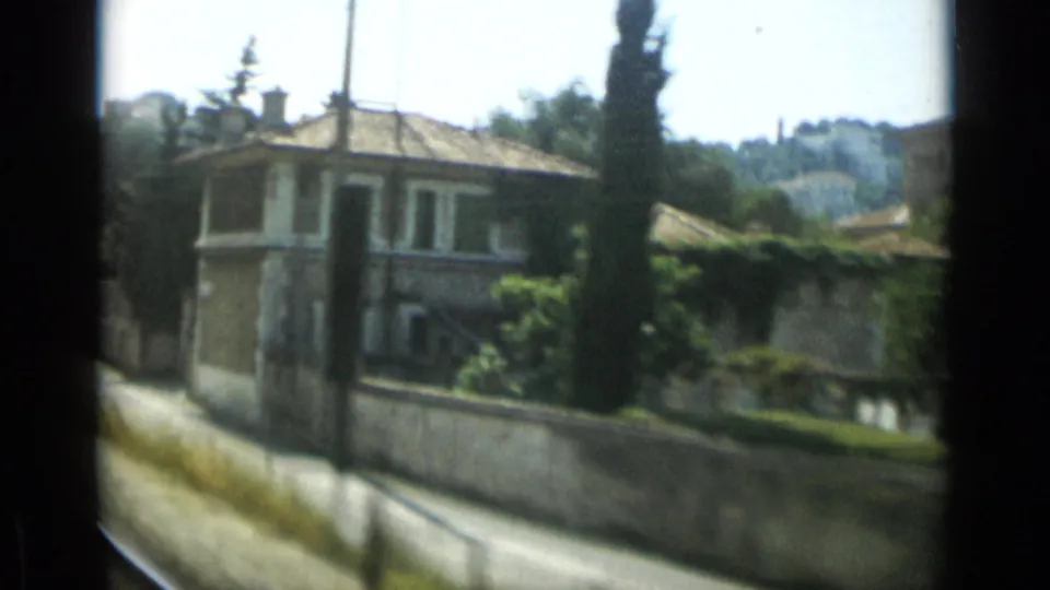 1962 Monaco roadside frame showing a tiled-roof villa, tall cypress, garden wall, and blurred road edge from a moving vehicle.
