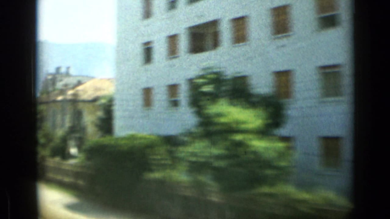 Blurred train-window view of a white apartment building, greenery, and nearby coastal town structures.