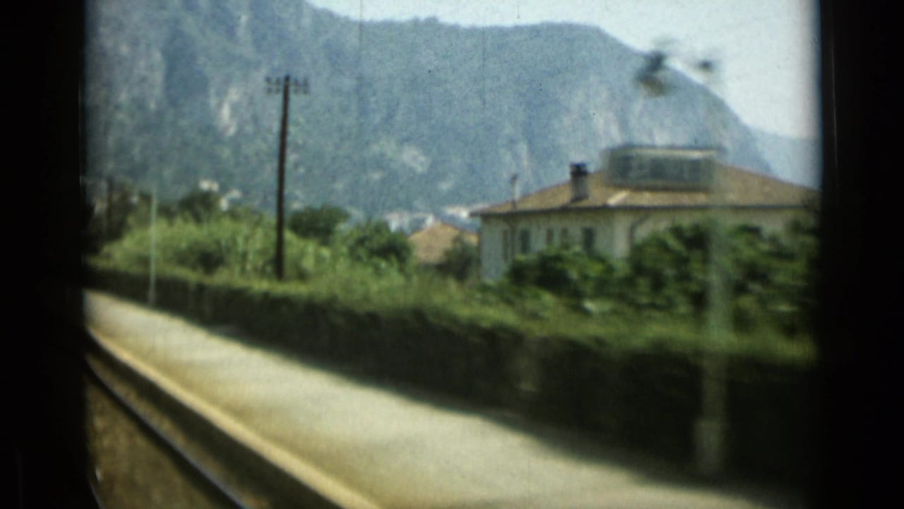 Rail platform beside tracks with a villa, greenery, utility pole, hillside, and sea in the distance.