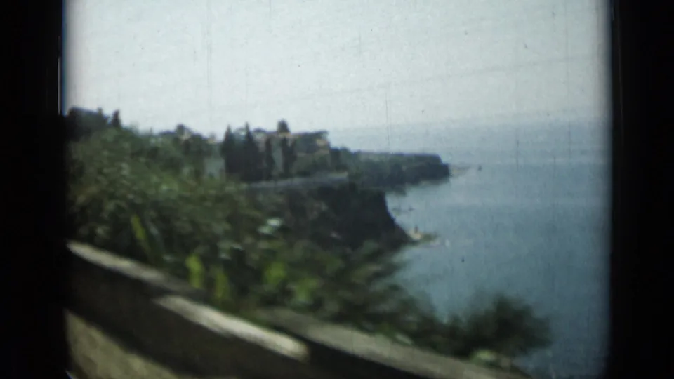 1962 Monaco train-window view of cliffside greenery, shoreline buildings, road edge, and Mediterranean water.
