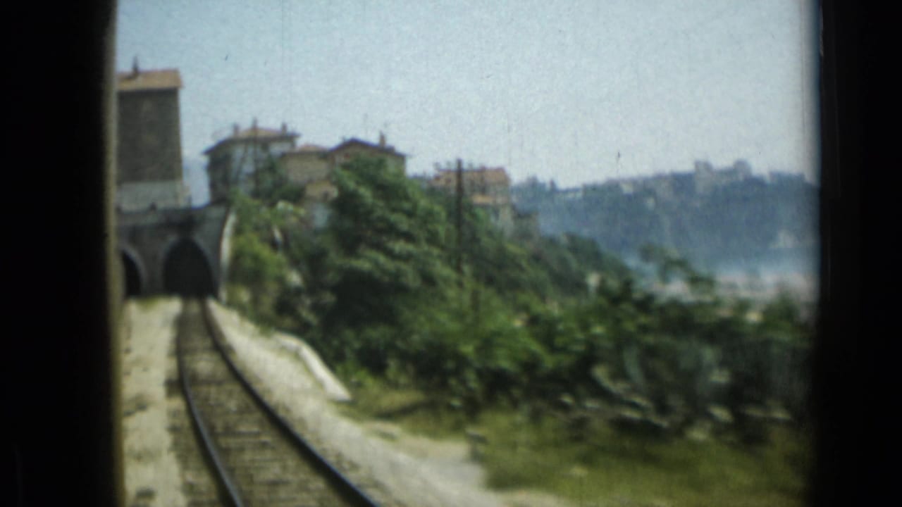 Rail tracks curve toward a stone tunnel beside trees and hillside buildings.