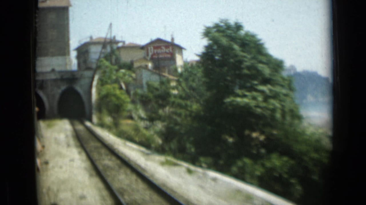Rail tracks run toward a tunnel with trees and a Prudel sign above nearby buildings.