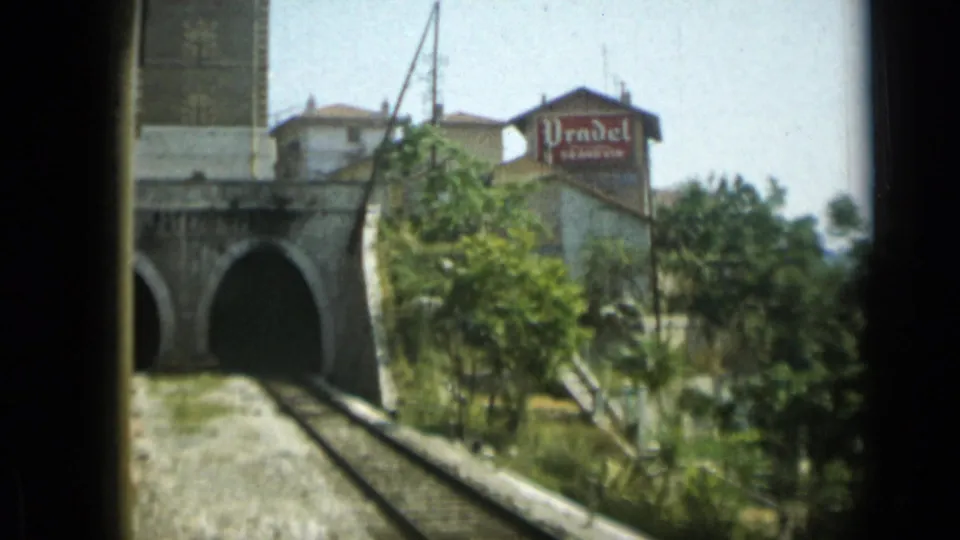 1962 Monaco 8mm frame showing the train approach closer to a stone tunnel, with trees, hillside structures, and a Prudel sign.