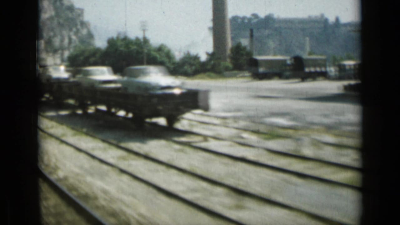 1962 Monaco rail view showing light-colored automobiles on a flatcar moving past tracks and industrial surroundings.