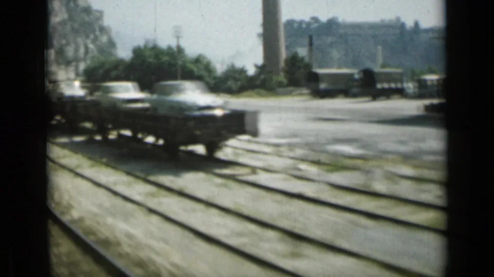 1962 Monaco rail view showing light-colored automobiles on a flatcar moving past tracks and industrial surroundings.