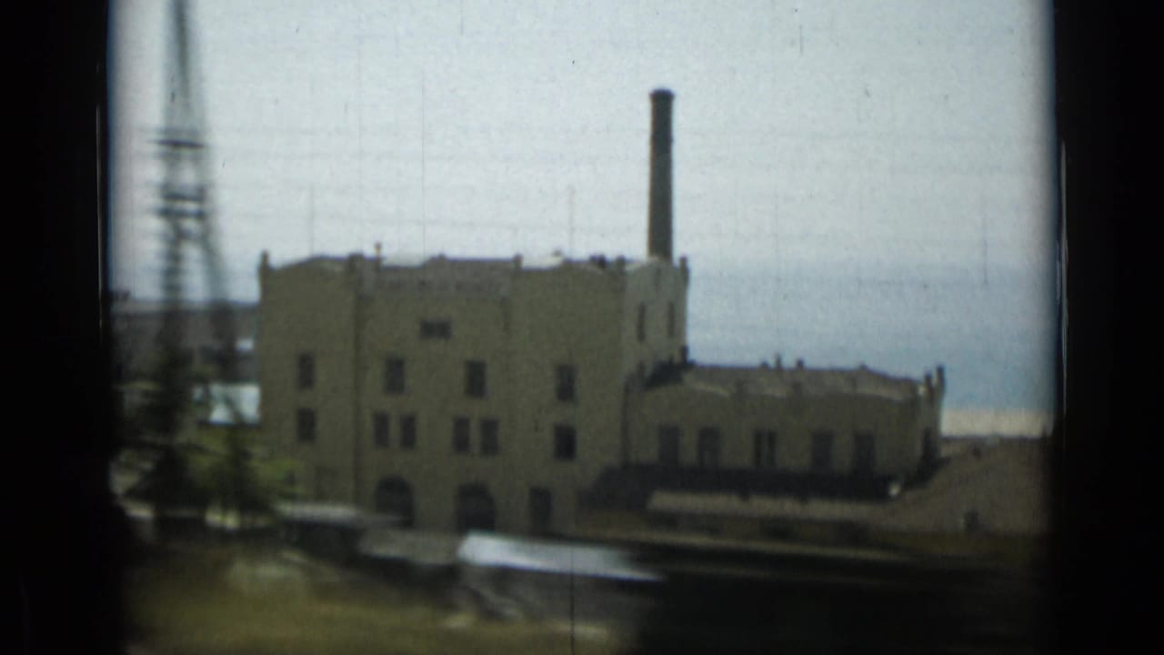 Car-window frame in Monaco showing a large pale industrial building, chimney, ocean background, and foreground motion blur.