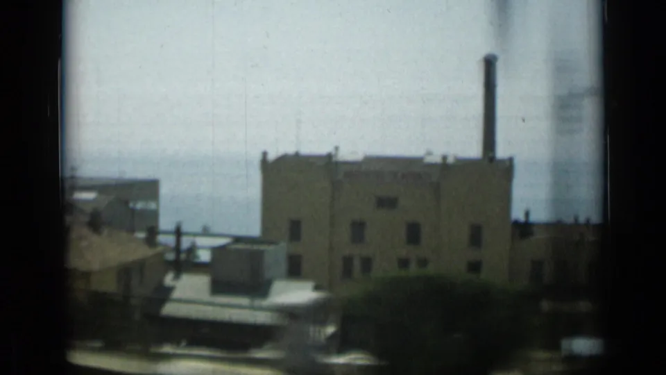 1962 Monaco view of a waterfront industrial structure with a chimney, foreground railway, and muted sea horizon.