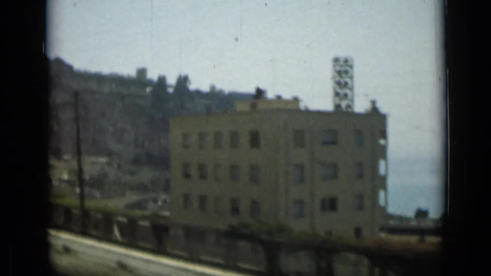 1962 Monaco home-movie frame showing a roadside building below a steep cliff, with pale sea visible in the background.