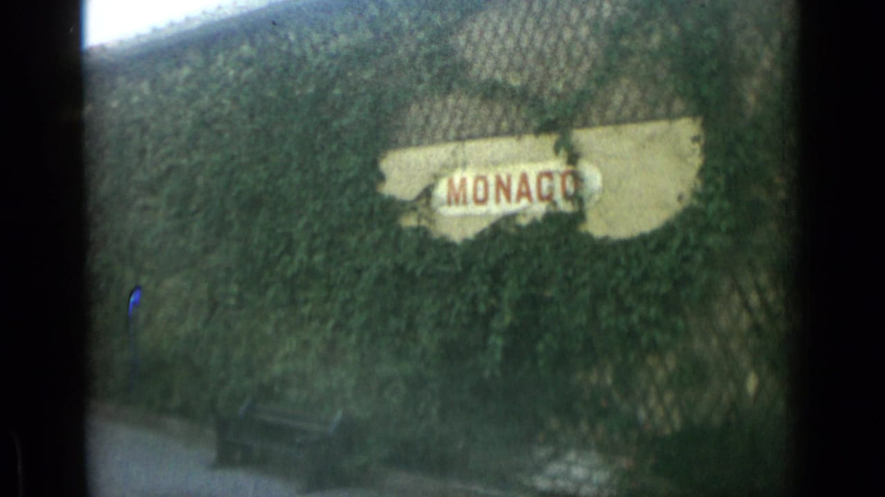 Ivy-covered MONACO sign seen through chain-link fencing with a bench below.