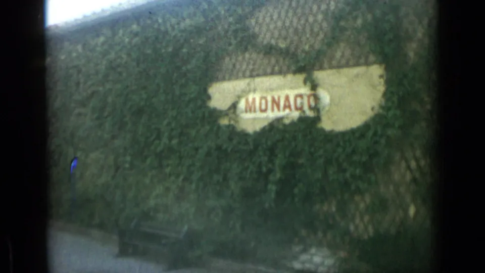1962 Monaco home-movie frame showing a weathered MONACO sign partly covered by ivy behind chain-link fencing, with a bench near the bottom.