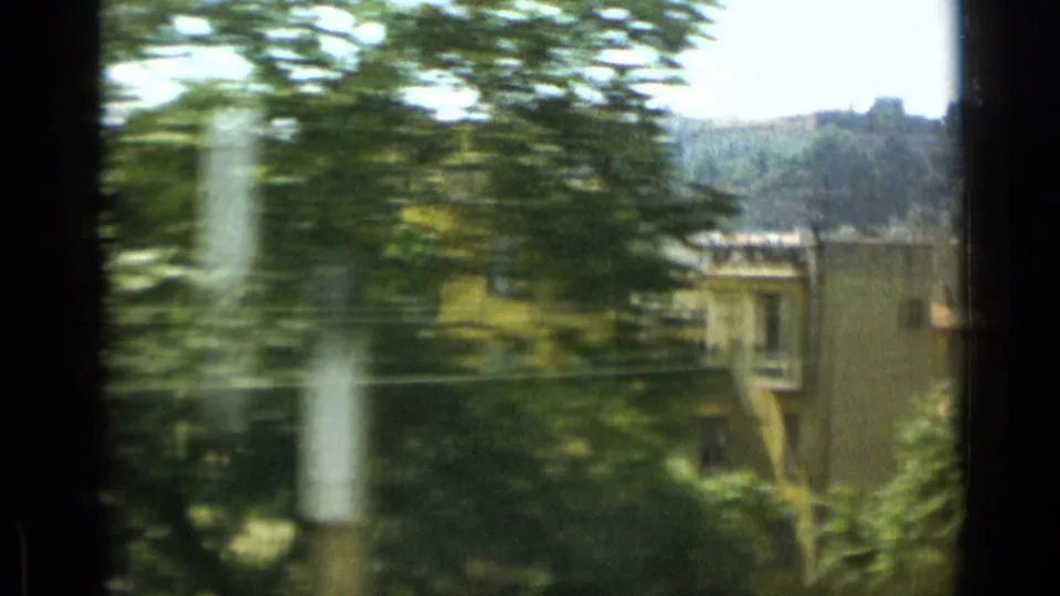 1962 Monaco travel frame showing pale buildings among green trees, with hillside terrain in the background and motion blur from handheld filming.