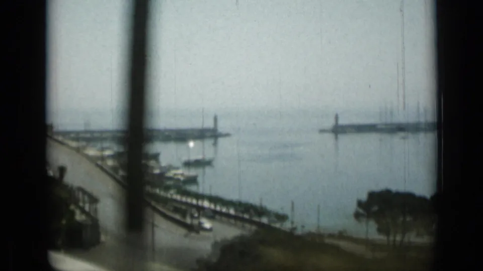 1962 Monaco harbor view with calm water, moored boats, quays, distant breakwaters, and vertical foreground obstruction from handheld framing.