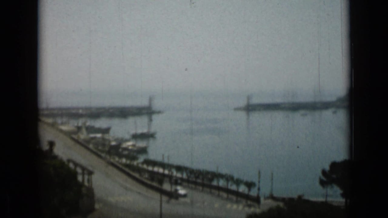 Hazy harbor view with breakwaters, waterfront road, trees, and dark film-frame edges.
