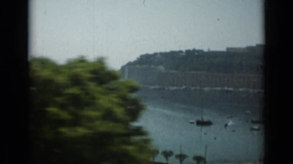 1962 Monaco marina frame with small boats on calm water, blurred roadside greenery, and a hazy hillside with shoreline structures beyond.
