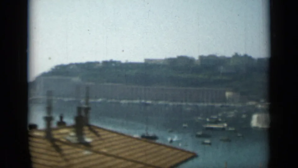 1962 Monaco harbor view with moored boats, a tiled roof and chimneys in the foreground, and hillside buildings across the water.