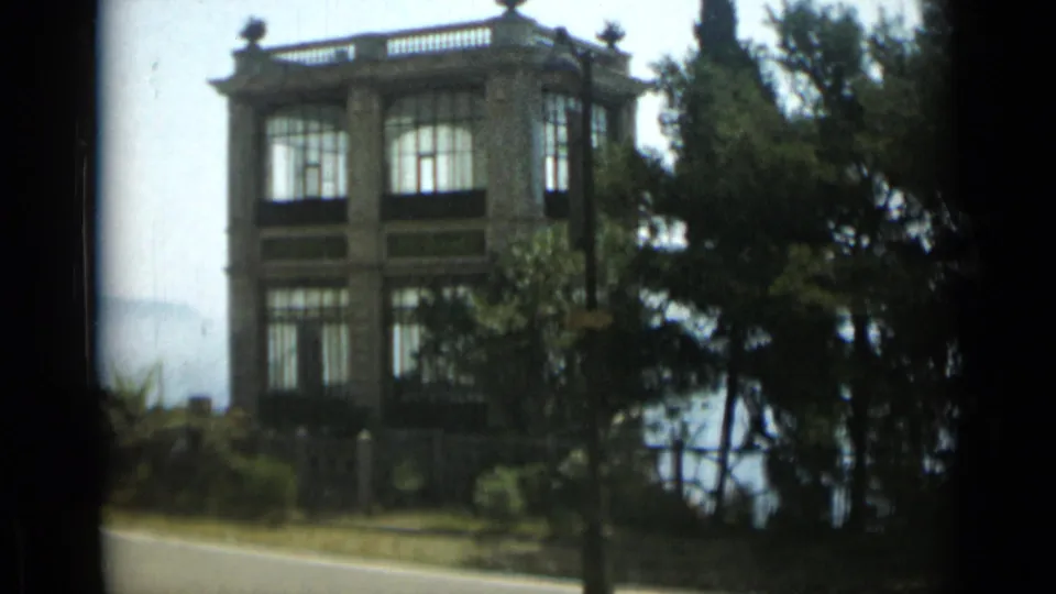 1962 Monaco waterfront scene with a grand stone building, tall windows, balcony details, trees, and water visible in the background.